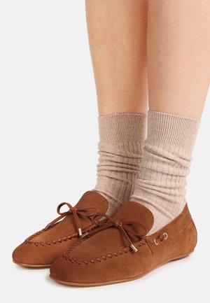Mocassins en suède marron avec lacets en cuir portés avec des chaussettes côtelées beiges sur des jambes nues, sur fond blanc.