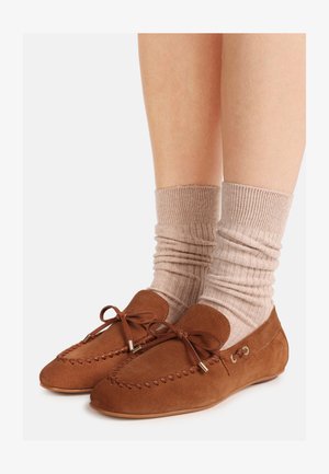 Mocassins en suède marron avec lacets en cuir portés avec des chaussettes côtelées beiges sur des jambes nues, sur fond blanc.