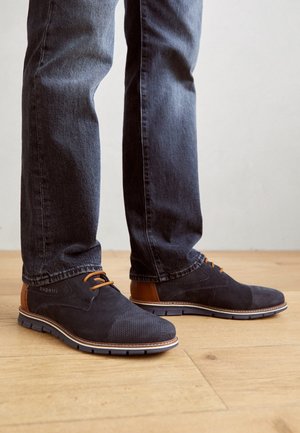 Homme portant des chaussures en daim bleu foncé à lacets avec des accents marron et un jean bleu foncé, debout sur un sol en bois.
