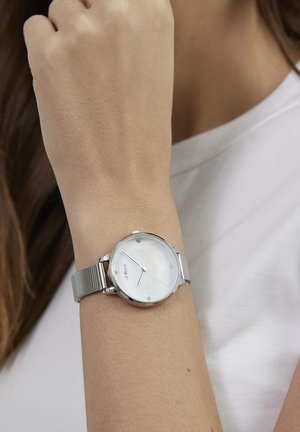 Silberne Mesh-Armband-Uhr mit rundem weißem Zifferblatt, das Stundenmarkierungen und ein minimalistisches Design zeigt. Die reflektierende Oberfläche verbessert die Sichtbarkeit.