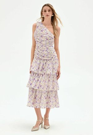 Femme portant une robe asymétrique à étages, fleurie lavande et blanche, avec des talons hauts dorés et des boucles d'oreilles créoles dorées.