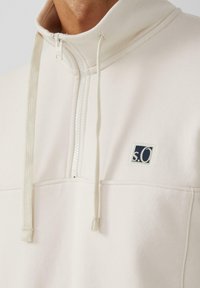 Sudadera de medio zip en color crema, hecha de una tela suave. Presenta un panel texturizado, cordones ajustables en la capucha y un pequeño parche con el logo en azul marino.