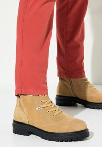 Botines de ante color mantequilla con suela de goma negra, ojales de metal y un cordón estiloso, combinados con pantalones rojos ajustados.