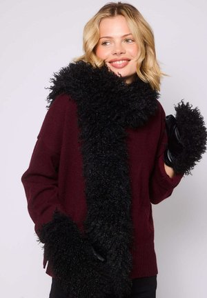 Donna sorridente che indossa un maglione bordeaux, una sciarpa fluffy nera e guanti fluffy neri abbinati su uno sfondo chiaro.