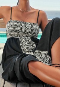 Schwarzes Kleid mit komplizierter weißer geometrischer Stickerei. Mit dünnen Trägern, einem enganliegenden Oberteil und einem fließenden Rock. Texturiertes Material.