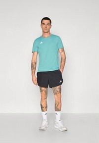 Tyrkysové sportovní tričko s krátkými rukávy, spojené s černými šortkami. Bílé značkové detaily na obou oděvech. Bílé tenisky a ponožky.