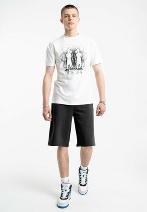 Młody mężczyzna w białej koszulce z nadrukiem przedstawiającym sylwetki i tekstem "ASTENSION", czarnych szortach do kolan, białych skarpetkach oraz sportowych butach z niebieskimi akcentami.