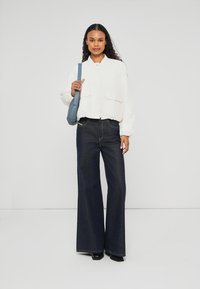 White cropped jacket with pockets, paired with dark blue wide-leg jeans. Model holds a blue shoulder bag. Smooth textures, simple design.