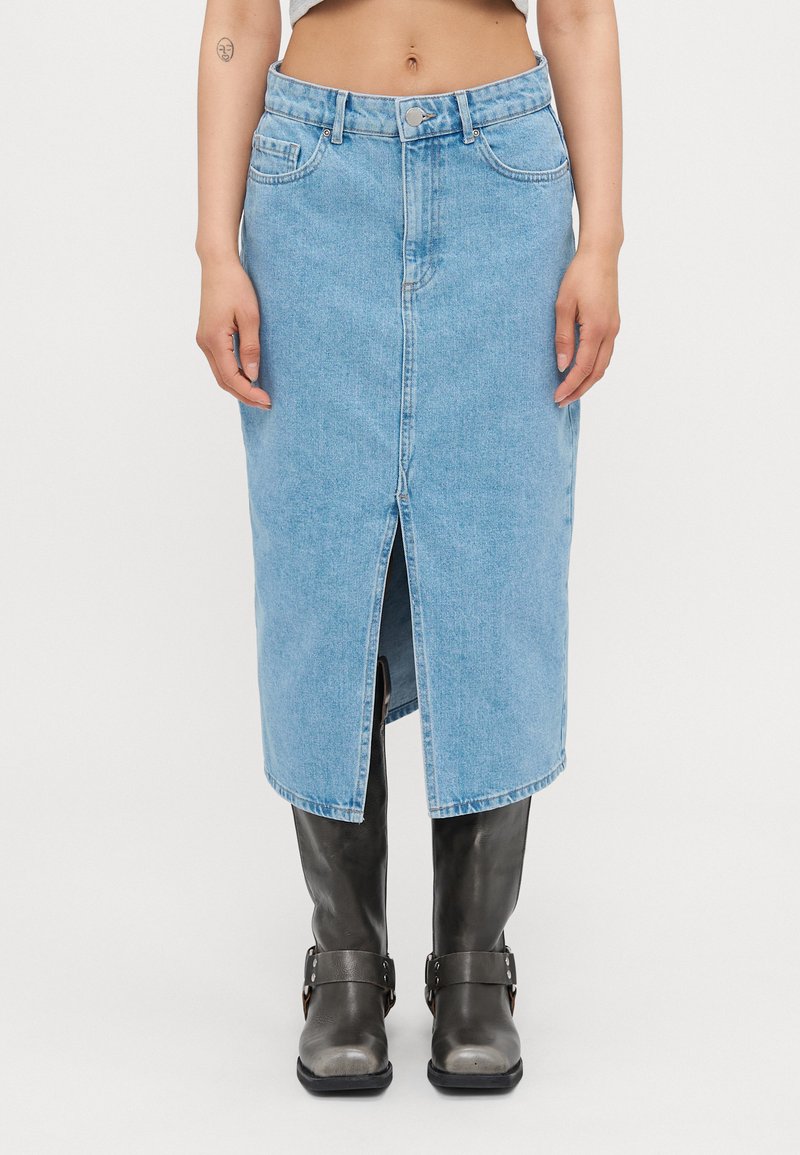 Saia midi em denim azul claro com fenda frontal, design de cinco bolsos e passantes para cinto. Usada com botas altas pretas até ao joelho. Textura suave.