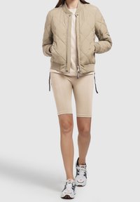 Pikowana kurtka typu bomber w kolorze khaki z prążkowanym obszyciem u dołu, zestawiona z beżowymi spodenkami kolarskimi. Model nosi białe i granatowe buty sportowe.