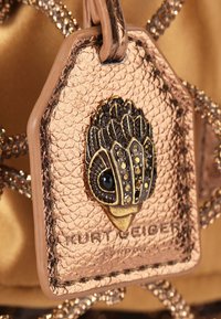 Roze gouden leren label met een gestructureerd oppervlak, voorzien van een gedetailleerd gouden en zwarte embleem en de geperste tekst "Kurt Geiger".