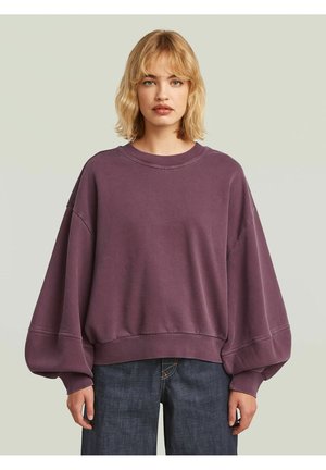 Kobieta o blond włosach ubrana w luźną fioletową bluzę z bufkami na rękawach i ciemnoniebieskie dżinsy na jednolitym jasnym tle.