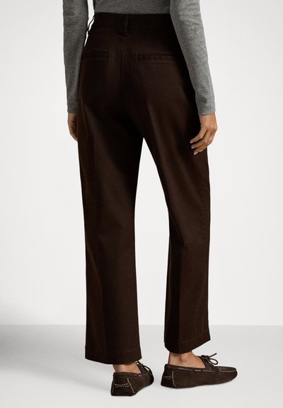 Άτομο που φοράει σκούρα καφέ παντελόνια με ευρύ πόδι και ταιριαστά σκούρα καφέ μοκασίνια, στέκεται μπροστά σε απλό φωτεινό φόντο.