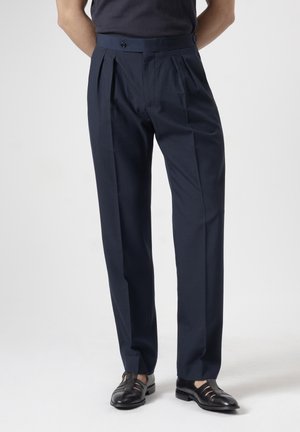 Hombre con pantalones de vestir azul marino plisados con cinturilla abotonada y zapatos de vestir de cuero negro sobre fondo liso.