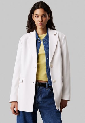 Blazer blanco con un corte entallado, que presenta un diseño de un solo botón, dos botones y bolsillos delanteros con solapa, superpuesto a una chaqueta de mezclilla y una blusa amarilla.