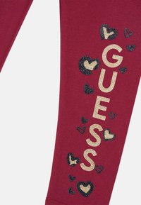Bordeaux leggings med guldglitter "GUESS" tekst, accentueret af sorte og guldglitter hjerter. Lavet af strækbart, blødt stof med en tætsiddende design.