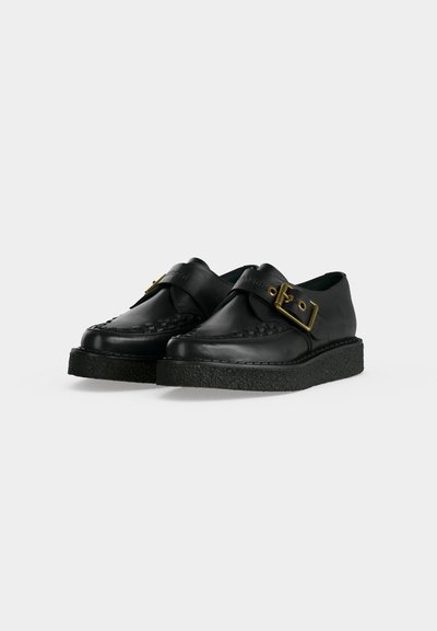 Μαύρα δερμάτινα loafers με χοντρές μαύρες λαστιχένιες σόλες, υφασμένα διακοσμητικά στο δάχτυλο και φαρδιά λουράκι με μεγάλη χρυσή αγκράφα στο πάνω μέρος.