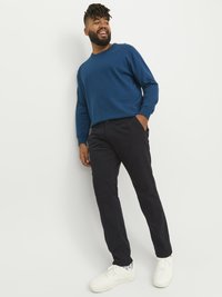 Blå långärmad tröja med ribbade muddars, kombinerad med mörka byxor och vita sneakers. Avslappnad outfit på en stående man med ansiktshår.