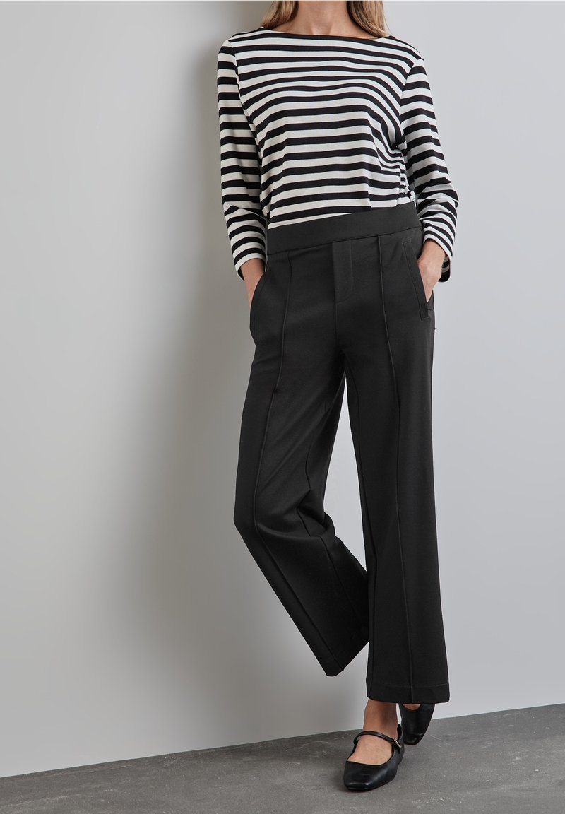 Femme debout les mains dans les poches, portant un pantalon noir à jambes larges, un haut rayé noir et blanc à manches longues et des chaussures plates noires.