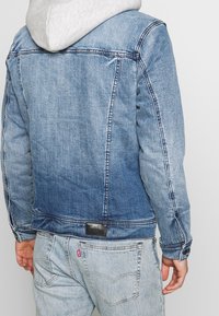 Veste en jean avec un dégradé de bleu, fermetures à boutons et coutures contrastantes. Sweat à capuche gris dépassant du col. Étiquette noire en bas.