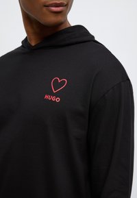 Μαύρο φούτερ με κουκούλα και απαλό υλικό, με κόκκινη καρδιά και τη λέξη "HUGO" με έντονα κόκκινα γράμματα στο στήθος.
