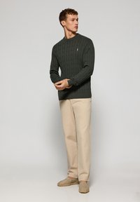 Grön stickad tröja med ett flätstickat mönster, rund halsringning, ribbade ärmavslut och nederkant. Matchad med beige byxor och avslappnade skor.