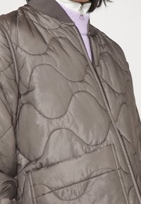 Gequilted jas in gedempt bruin, met een ritssluiting aan de voorkant, ronde kraag en grote opgestikte zak. Getextureerd golfpatroon voegt diepte toe.