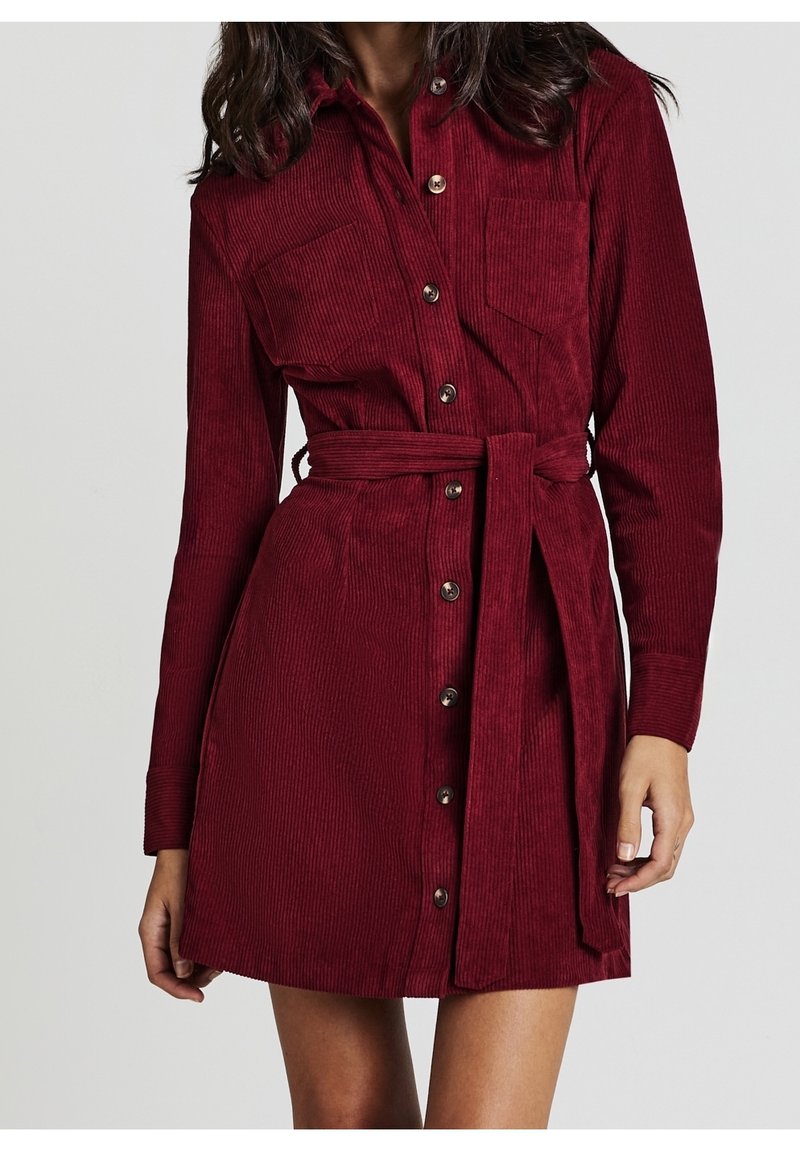 Femme portant une robe en velours côtelé bordeaux à manches longues, boutonnée sur le devant, avec des poches poitrine et une ceinture assortie nouée à la taille.