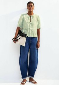 Groen en witte gestreepte peplumtop met keyhole-detail, gecombineerd met losse blauwe spijkerbroek en platte witte sandalen, terwijl ze een zwart-beige tas vasthoudt.
