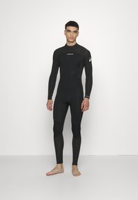Mute nero con maniche lunghe, superficie strutturata, design attillato, toppe per le ginocchia e un logo sul braccio sinistro. Il modello è a piedi nudi.