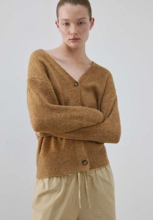 Joven con piel clara y cabello recogido en un moño, lleva un cárdigan marrón abotonado y pantalones beige con cuadros, de pie con los brazos cruzados.