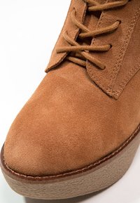 Brązowy zamszowy botek za kostkę z okrągłym noskiem, sznurowany z przodu, z kremową, teksturowaną platformą. Gładka cholewka z przeszyciami jako akcentami.