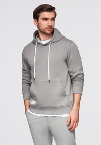 Šedá mikina s klokaní kapsou, bílými šňůrkami a látkovým štítkem. Měkká textura, volný střih, nošena přes bílou košili.