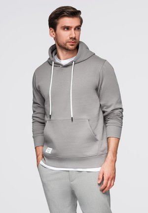 Felpa grigia con tasca a canguro, cordini bianchi e etichetta in tessuto. Tessuto morbido, vestibilità rilassata, indossata sopra una maglietta bianca.