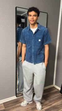 Νεαρός άνδρας στέκεται με τα χέρια στις τσέπες, φορά μπλε πουκάμισο με κοντά μανίκια, γκρι φόρμα και λευκά αθλητικά παπούτσια μπροστά σε καθρέφτη τοίχου.