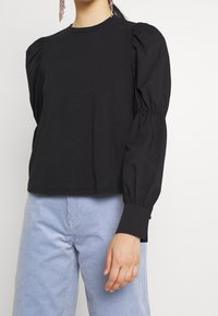 Donna che indossa una maglietta nera a maniche lunghe con spalline a sbuffo e polsini arricciati, abbinata a pantaloni in velluto a coste blu chiaro.