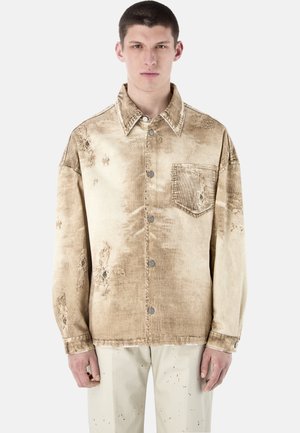 Młody mężczyzna w beżowej, postarzanej kurtce sztruksowej z metalowymi guzikami i kieszenią na piersi, stojący na tle jednolitego, jasnego tła.