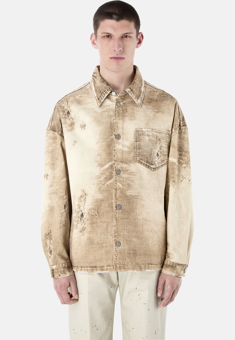 Jeune homme portant une veste en velours côtelé beige usée avec des boutons en métal et une poche poitrine, debout devant un fond clair uni.