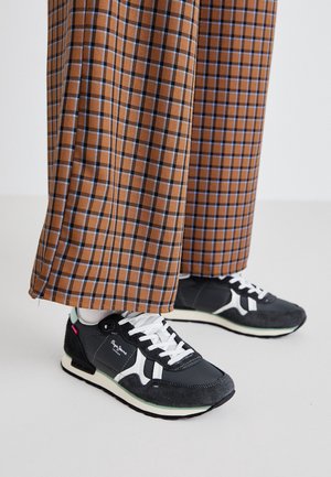 Schwarz-weiße Sneaker getragen zu braunen, karrierten, weit geschnittenen Hosen vor einem schlichten, hellen Hintergrund.