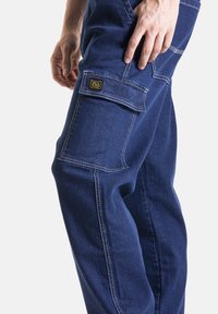 Salopette en denim bleu foncé avec des coutures blanches et une poche latérale. Comprend une étiquette de marque visible près de la poche. Coupe décontractée.