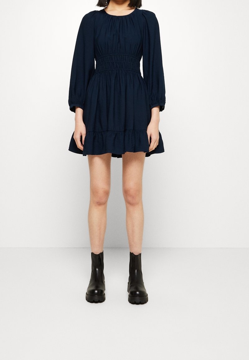 Robe bleu marine à manches longues, taille froncée et ourlet volanté. Associée à des bottines noires à la cheville. Tissu lisse, design simple.