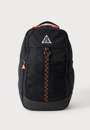 Sac à dos Nike ACG noir avec poche avant en mesh, tirettes de fermeture éclair orange et sangle cousue en zigzag orange sur fond uni.