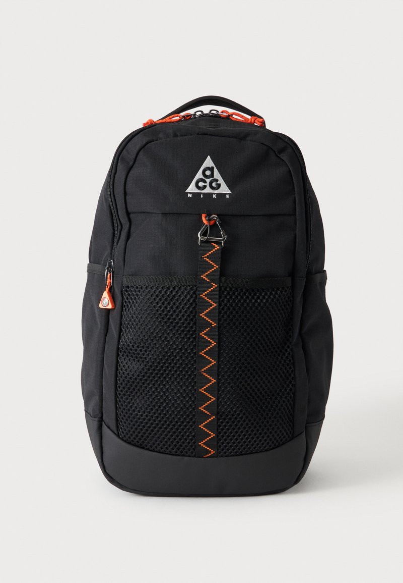 Sac à dos Nike ACG noir avec poche avant en mesh, tirettes de fermeture éclair orange et sangle cousue en zigzag orange sur fond uni.