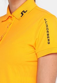 Gul polo skjorta med krage, med knappar och svart logodetalj på kragen och ärmen. Tillverkad av ett mjukt, lätt tyg.