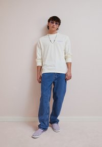 Long-sleeve cream shirt with purple lettering, blue denim jeans, and purple checkered slip-on shoes. Silver chain necklace and sunglasses.