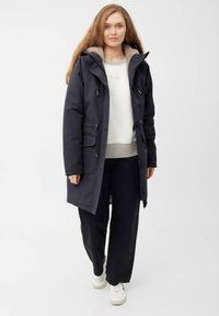 Námořnická parka s kapucí podšitou kožešinou, předními kapsami a nastavitelnými šňůrkami. Nositá přes šedou mikinu a černé kalhoty, doplněná bílými teniskami.