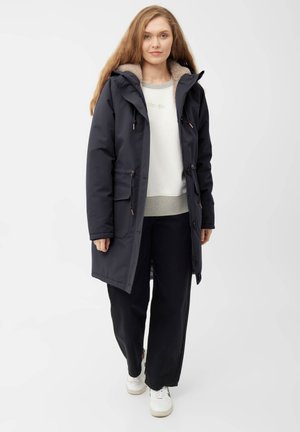 Marine parkajack met een met bont gevoerde capuchon, voorkezen en verstelbare toggles. Draag het over een grijs sweatshirt en zwarte broek, gecombineerd met witte sneakers.