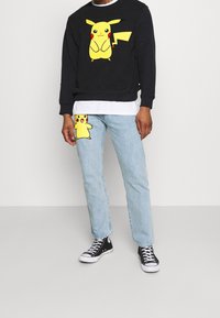Felpa nera con una grande grafica di Pikachu gialla, abbinata a jeans azzurri chiari con una piccola toppa di Pikachu. Sneakers nere.