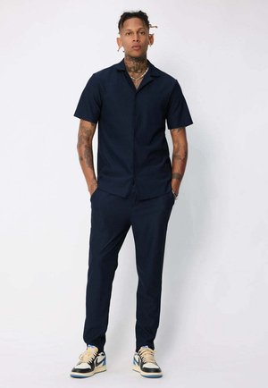 Chemise à manches courtes bleu marine à boutons, pantalon fuselé assorti et baskets beiges avec accents bleus ; tatouages visibles sur les bras.