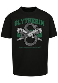Melna kokvilnas T-kreklu ar Slytherin dizainu, uz kura ir attēlots čūska un šķērsoti burvju nūjas. Zaļā krāsā ir uzraksti "SLYTHERIN" un "HOGWARTS BURVJU SKOLA."